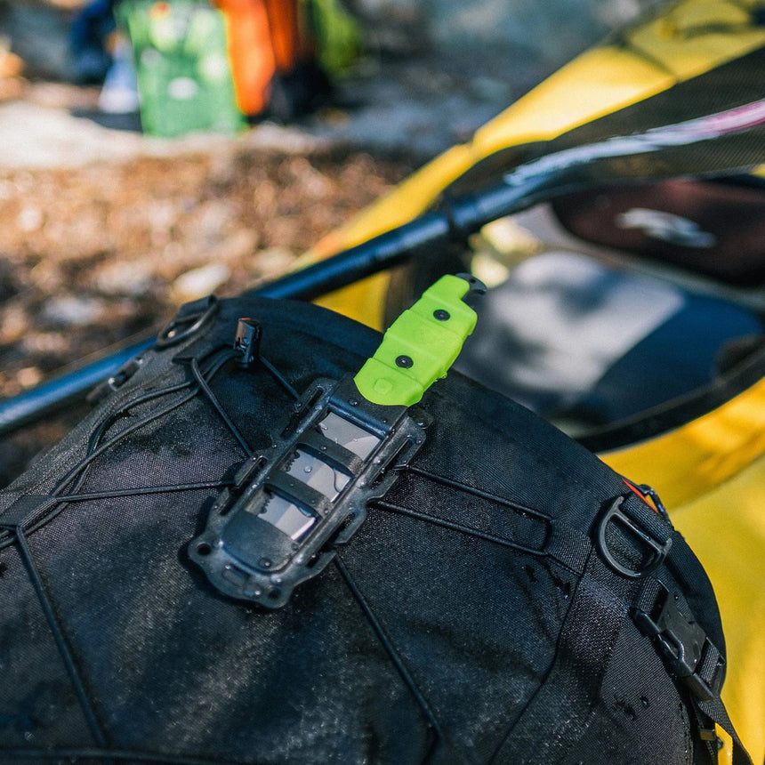 Green Gear Aid Akua River Knife Attached to the outside of a black Kayaking Gear Bag with a yellow kayak and paddle in the background