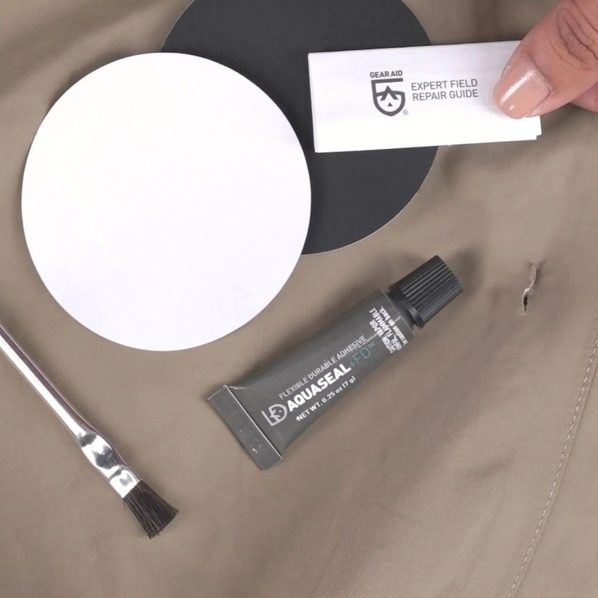 Gear Aid Aquasure +FD Repair Kit laid out on fabric showing contents : an expert field repair guide, an adhesive application brush, Aquasure +FD Adhesive, a clear and a black tenacious tape patch
