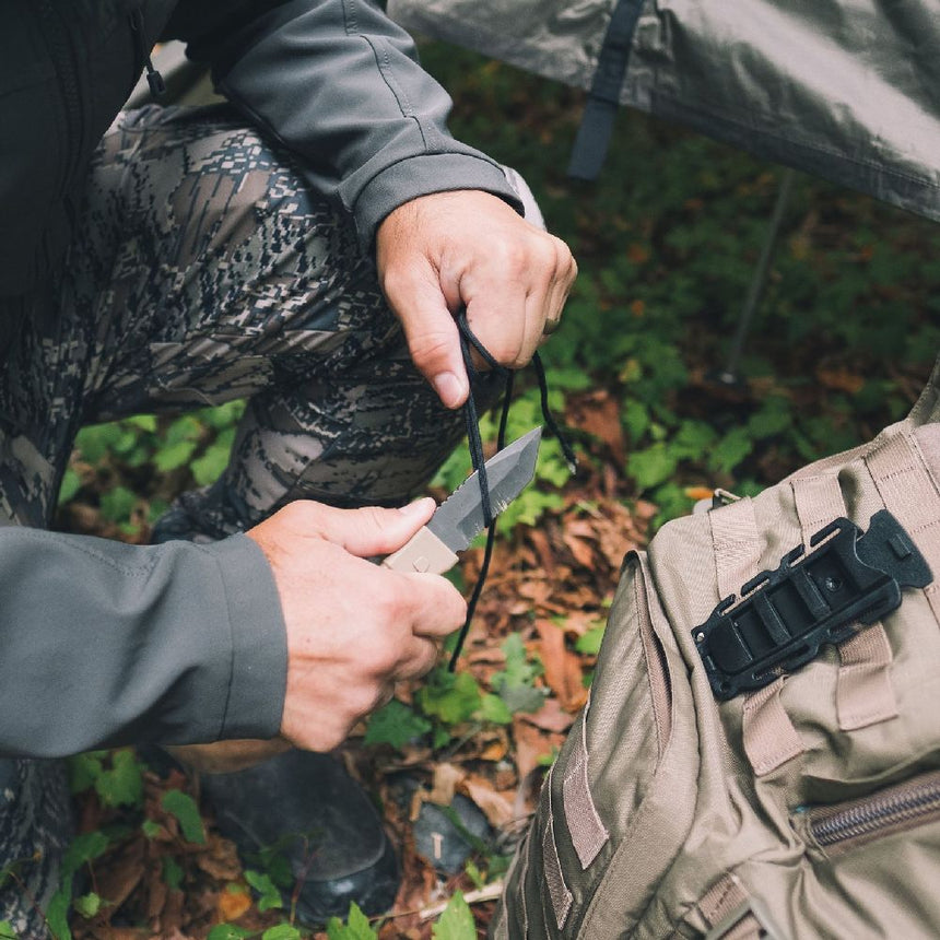 Model using a coyote coloured Gear Aid Kotu knife cutting a cord outside beside a gear bag with the quick release sheath attached