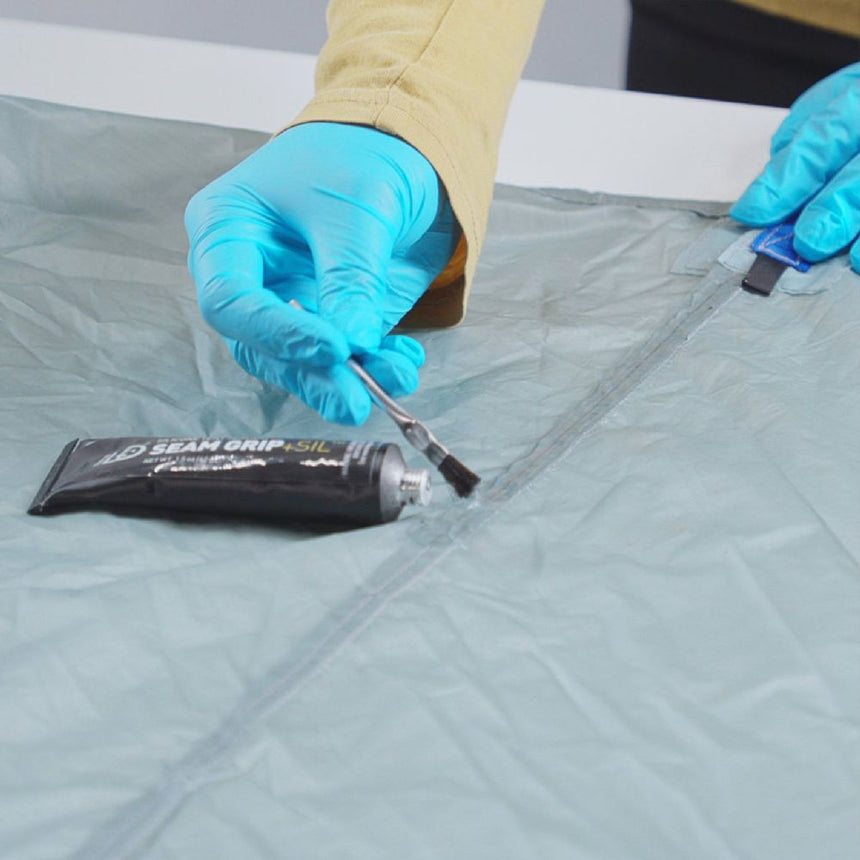 User wearing protective blue gloves applying Gear Aid Seam Grip+ SIL Silicone Tent Sealant with a small brush on the seam of a grey tent
