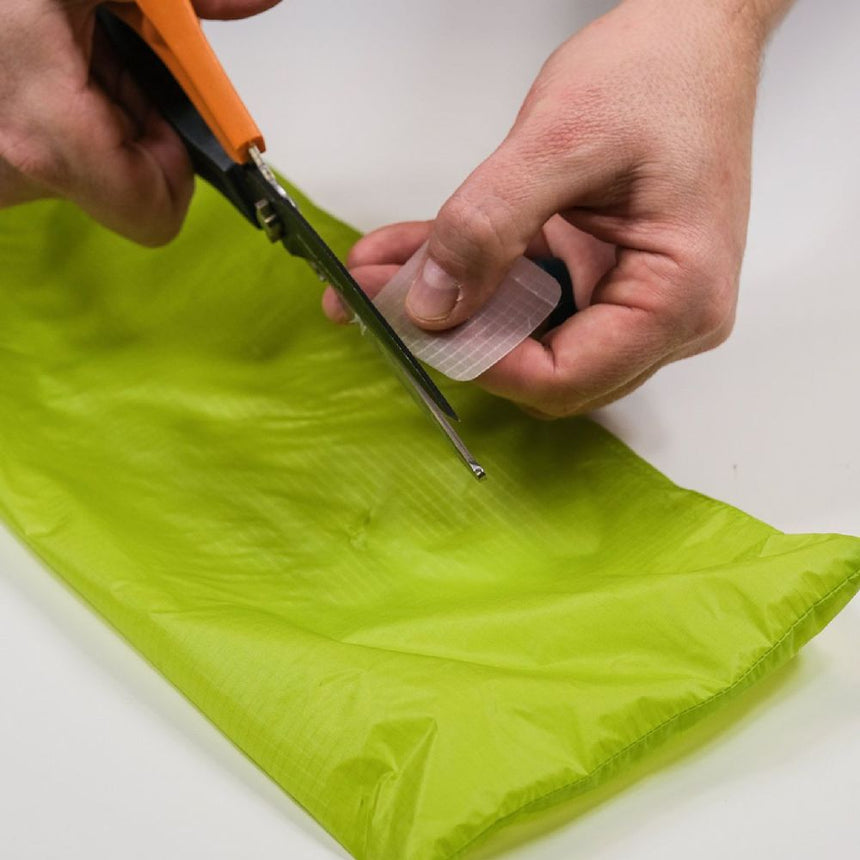 Hand model using a scissors to cut Semi Transparent Peel and Stick Gear Aid Tenacious Tape Sil Nylon Repair Patch to apply to a tear in an ultralight stuff bag for a jacket