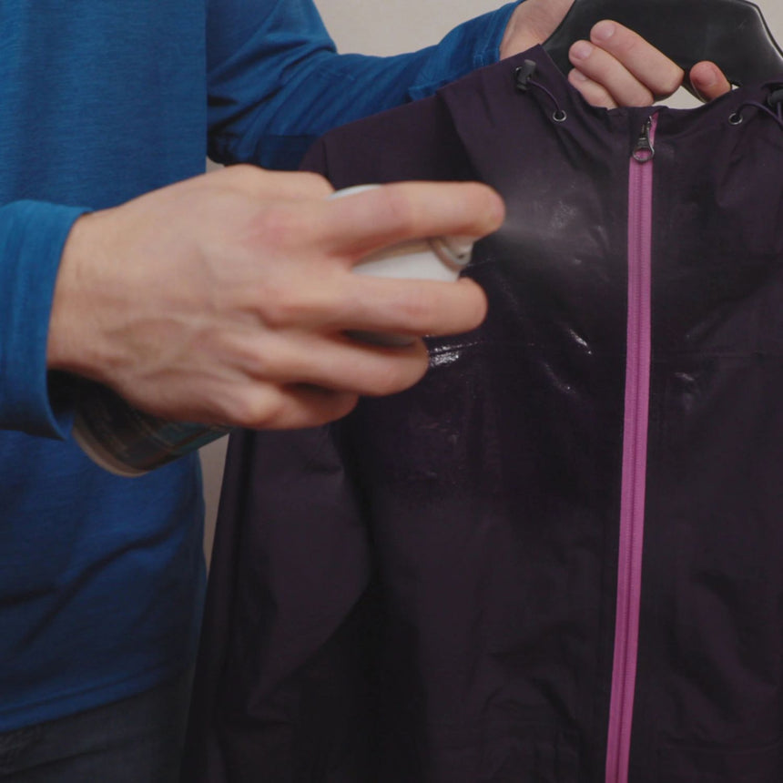 Person holding a purple jacket on a coat hanger and spraying Gearaid Water Repellent Spray on the jacket