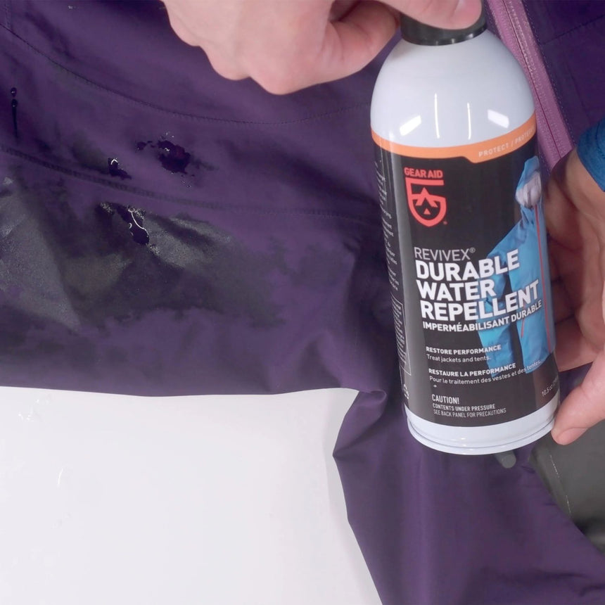 Close up of hands holding Gearaid Durable Water Repellent Bottle beside sprayed purple jacket