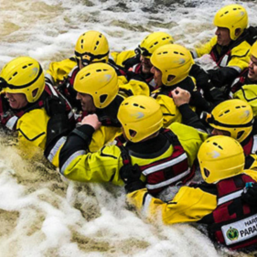 A group of rescue professionals wearing yellow helmets and suits linked together in a swift water environment, demonstrating teamwork and safety measures during a rescue scenario.