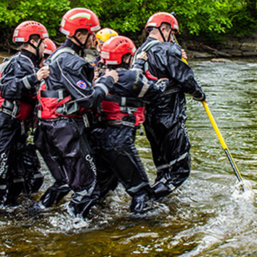 Rescue personnel wearing red helmets and waterproof suits using the Reach and Rescue Wading Pole to navigate through shallow, turbulent water during a rescue operation.