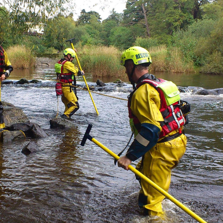 Two rescue workers equipped with yellow helmets and waterproof gear crossing a rocky riverbed using the Reach and Rescue Wading Pole for support and stability.