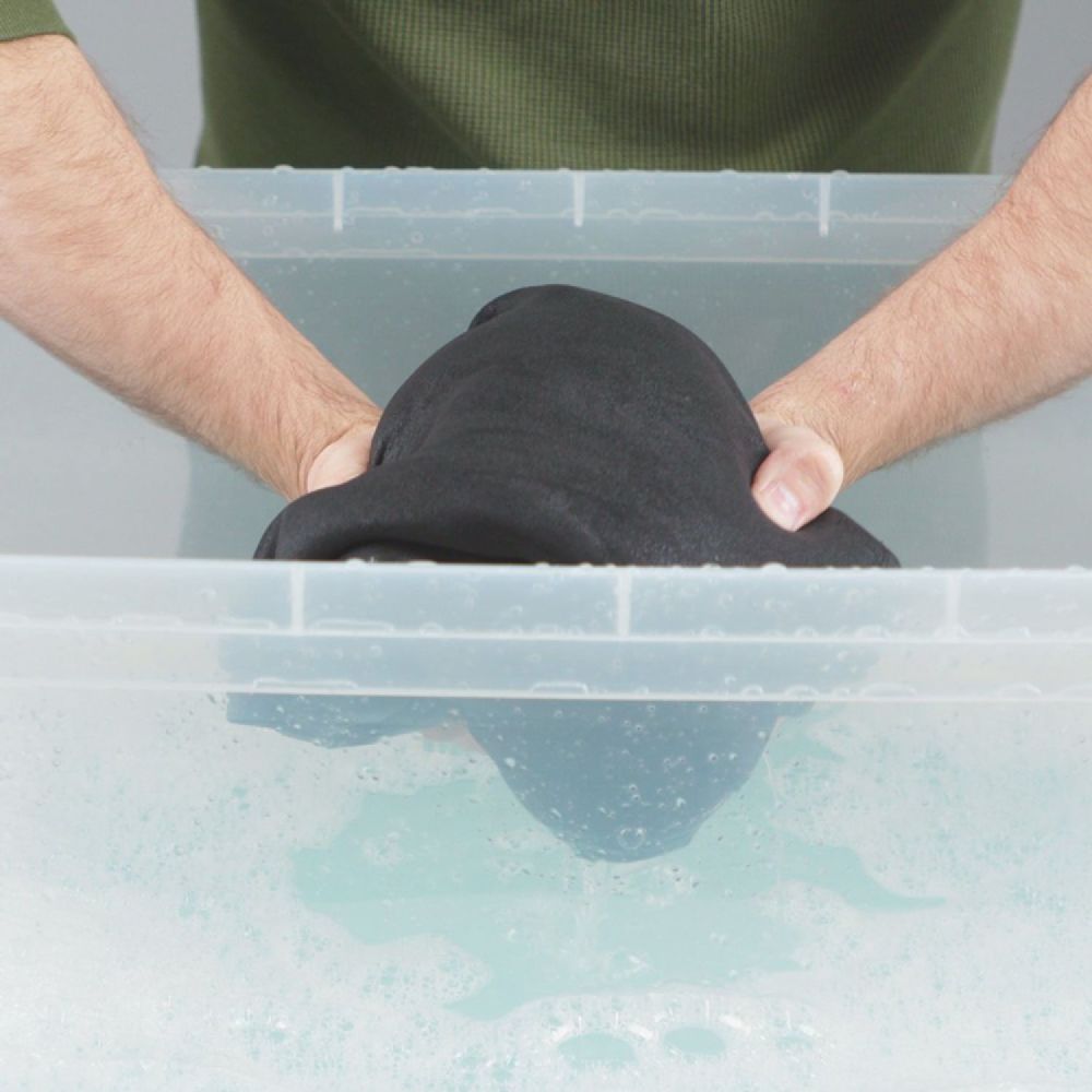 Hand model putting neoprene suit in a transparent basin with Gear Aid Revivex Wetsuit Shampoo diluted in water