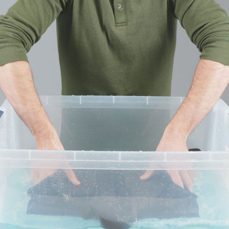 Hand model about to hand wash neoprene suit in a transparent basin with Gear Aid Revivex Wetsuit Shampoo diluted in water